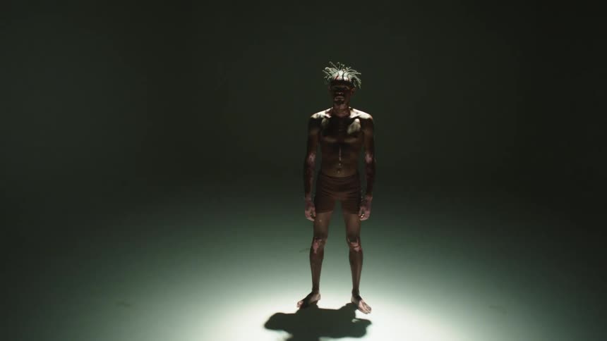 A man wearing only shorts stands under a spotlight in a room
