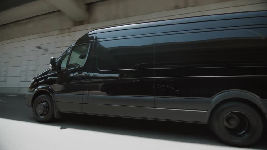 A black transport van travels along a highway beneath a bridge during the day