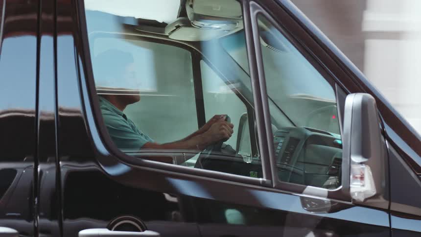 A focused working man carefully drives a black transport van along a highway during the day