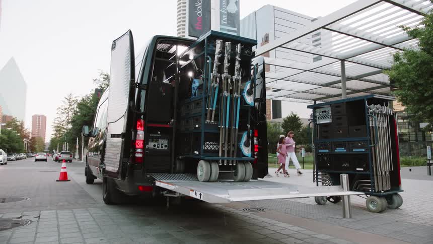 A man pushes a cart with equipment out of a van near a building entrance with people walking by