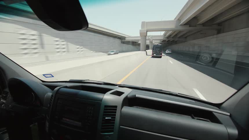 Interior passenger view through the windshield of a black transport van traveling on a divided highway on a bright sunny day