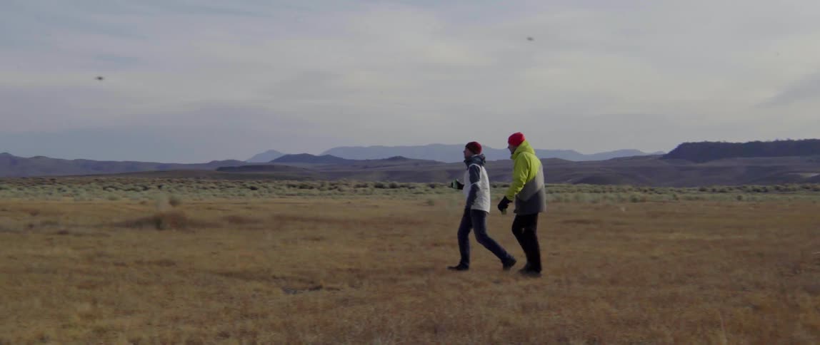 Two men in heavy coats walk across a field of short brown grass