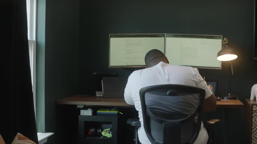 A man sits at a computer desk in front of two monitors
