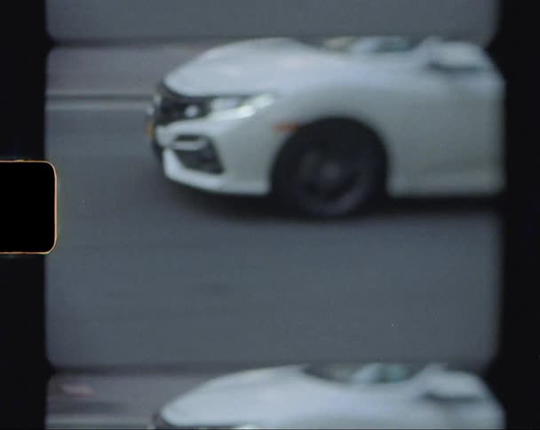 A white sedan drives along a city street lined with trees and pedestrians