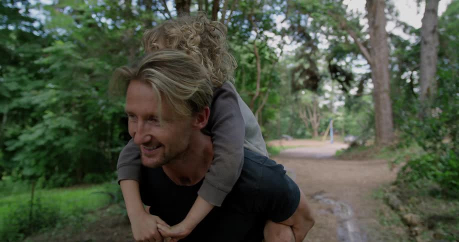 A father carries his son on his back through a forest trail