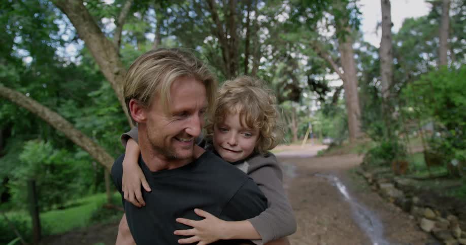 A man laughs and talks as he gives his son a piggy back along a path