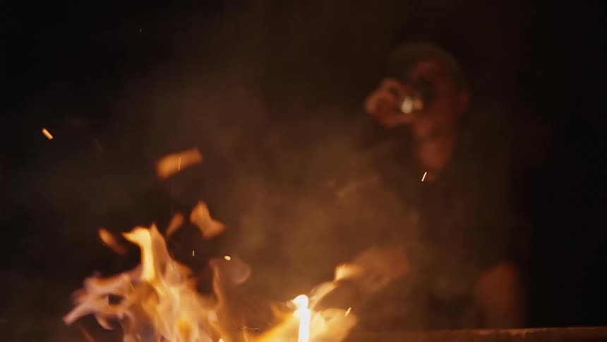 A man is sitting outside next to a bonfire and having a drink as he watches the flames