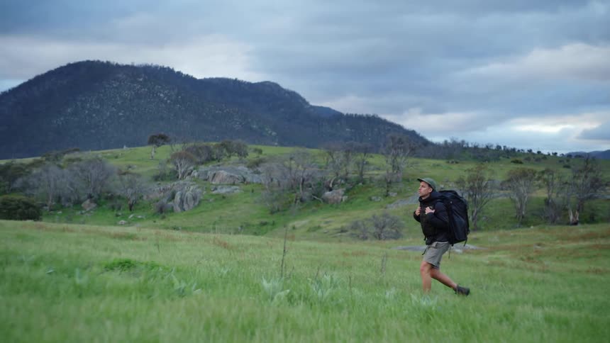 A man wearing shorts and a big backpack is walking across the grass with mountains in the distance