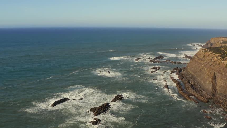 Waves foam as they crash against rocks near shore and cliff under blue sky