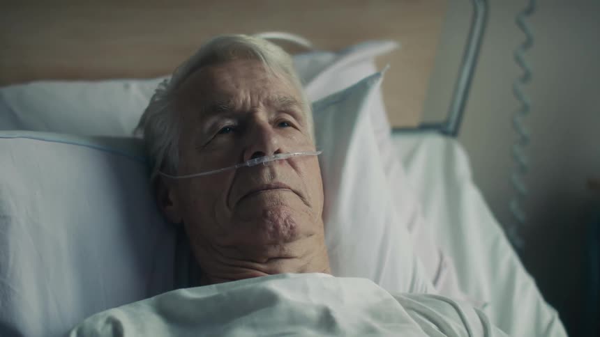 A elderly man lying in a hospital bed nods his head and speaks