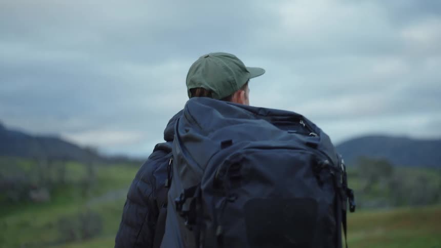 A man hikes through a grassland while wearing a large backpack on a cloudy day