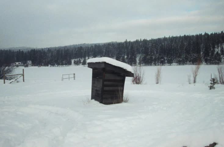 A snow covered field and outhouse with a forest of trees in the distance
