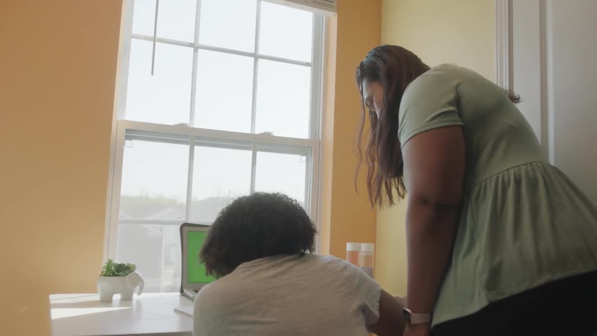 A mother helping her daughter study at the table next to a window inside the house
