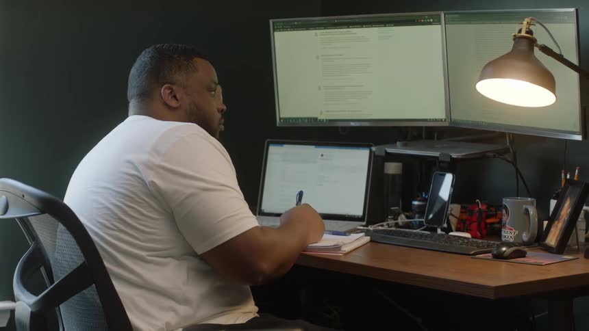 A man writes in a notebook as he sits at a desk covered in computer equipment