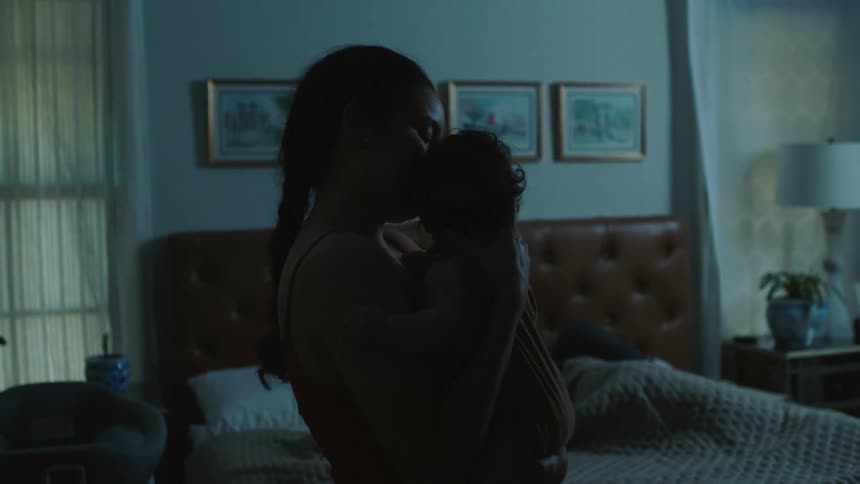 A woman rocks a baby in her arms while standing in a dimly lit bedroom as her partner sleeps on the bed behind her