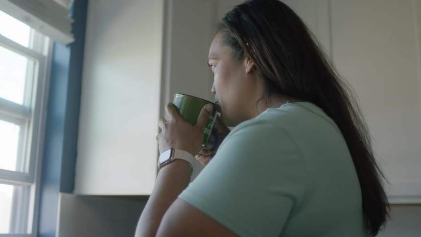 Woman standing at her kitchen looking through the window while drinking a coffee
