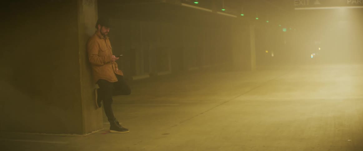 Man wearing baseball cap reclined leisurely against a column inside a foggy parking garage