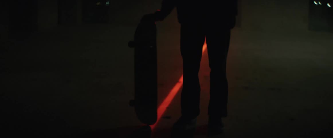 A man stands on a road at night and spins his skateboard around
