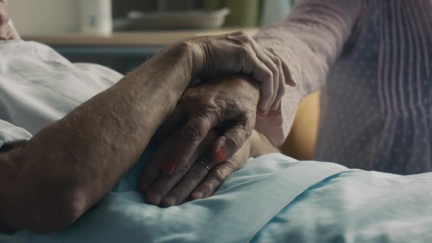 Old man laying at a hospital bed while his wife gently touches his hand