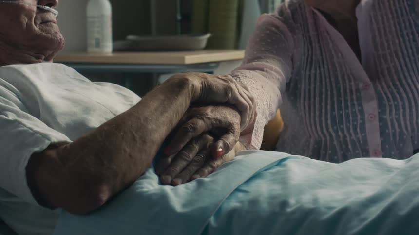 An elderly woman reaches her hand out to an elderly man laying in a hospital bed