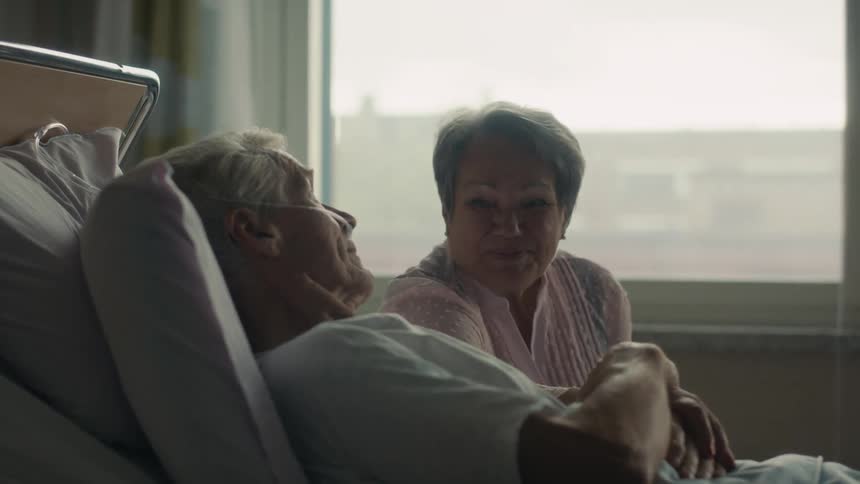 A woman reaches out to grab the hand of an elderly man laying in a hospital bed and they both smile at each other