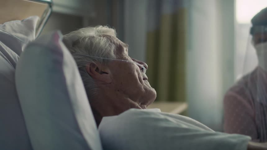 A man lays in a hospital bed and looks at a woman sitting beside him