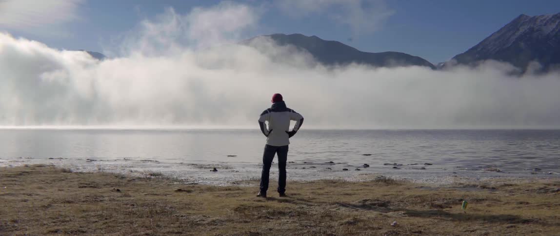Man Standing Near the Water by a Mountain Taking in the Quiet Vast Landscape.
