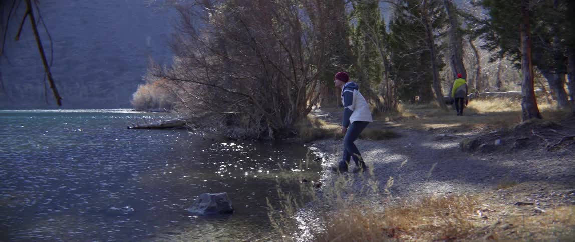A man walks down the river bank and puts his hand in the water
