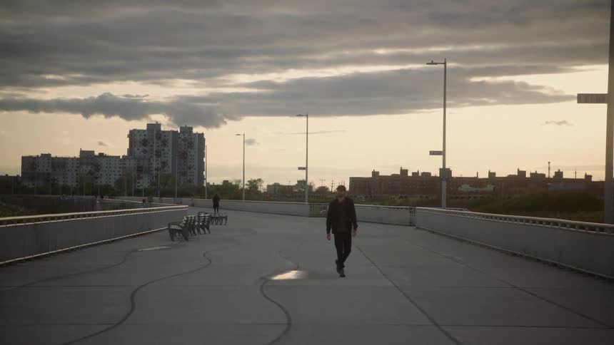 A man wearing dark colored clothes is walking on a concrete walkway with buildings in the distance under a cloudy sky