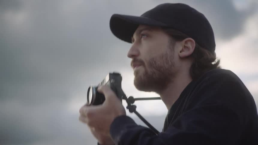 A man adjusts his camera lens before raising the camera to his face to take a photograph while standing outdoors during the day