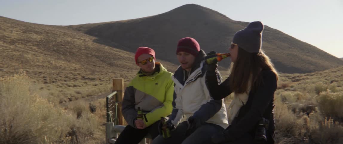 Three people sitting by hill drinking and talking with relaxed friendship vibe.