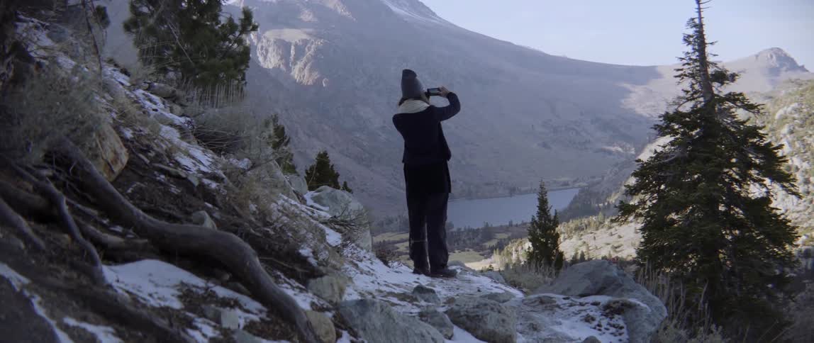 A woman stands on some rocks on a mountain range taking pictures