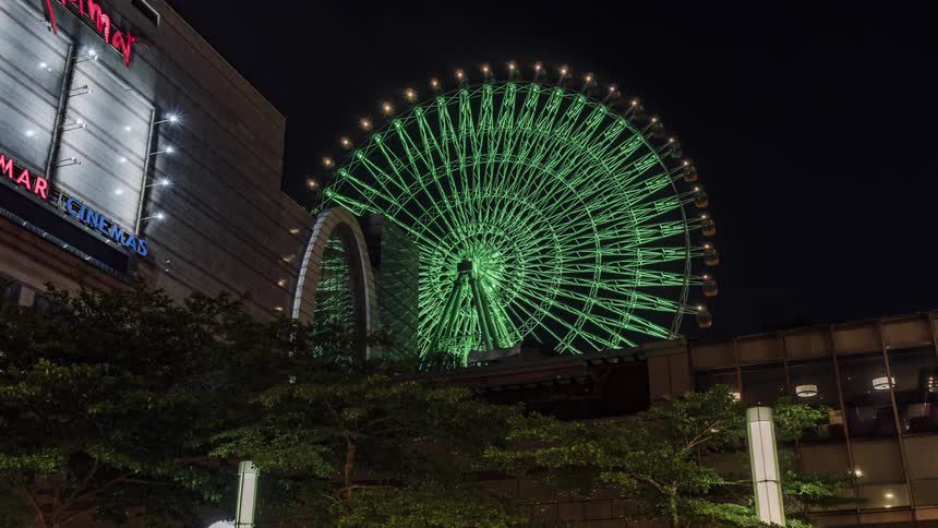 Timelapse footage of a green ferris wheel turning in behind a mall at night