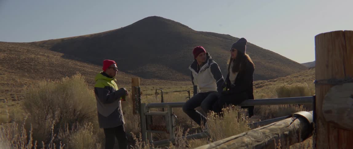 Three People Talking by a Fence Beside Gentle Hills in a Warm Outdoor Conversation.