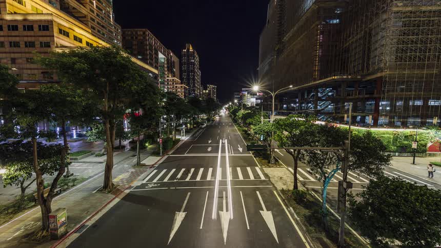 Headlights blur as traffic moves on a wide city street with pedestrians crossing