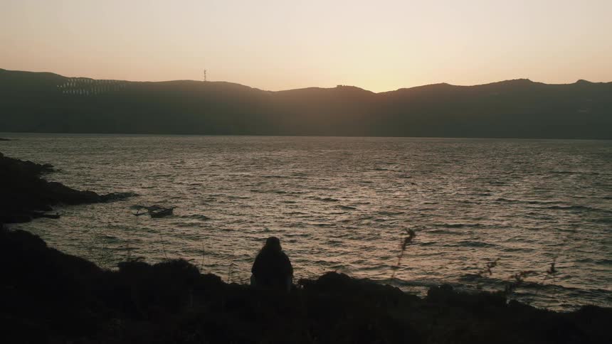 Someone is sitting near the lake as plants move in the breeze as the water ripples and a silhouette of mountains is along the shoreline