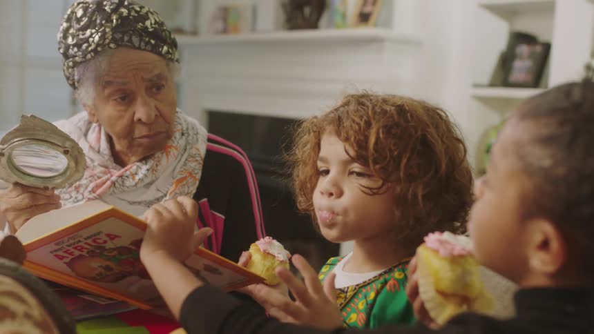Grandma uses a magnifying glass to read a book about africa to her grandchildren while they eat cupcakes