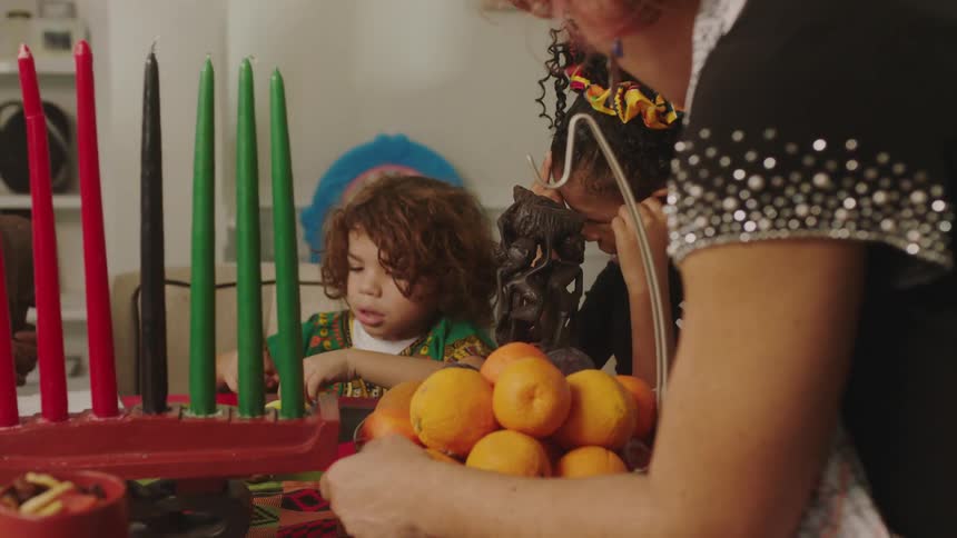 Mom places a bowl of fruit on the table where grandma is talking to the children about kwanzaa