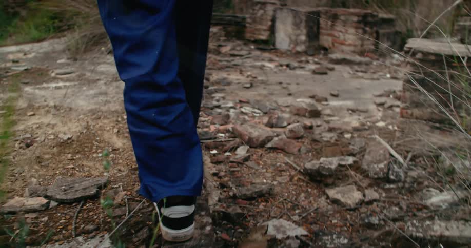 A person walking through debris stones and tall grass onto what remains of a brick wall