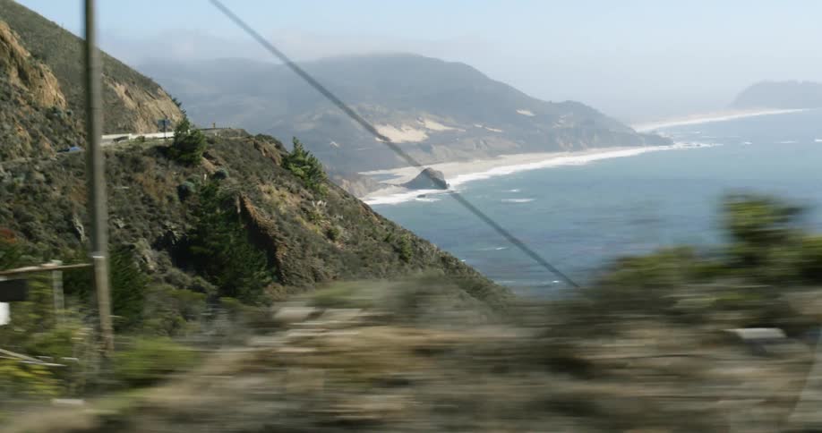 Cars move along highway hugging mountain and overlooking foggy shore