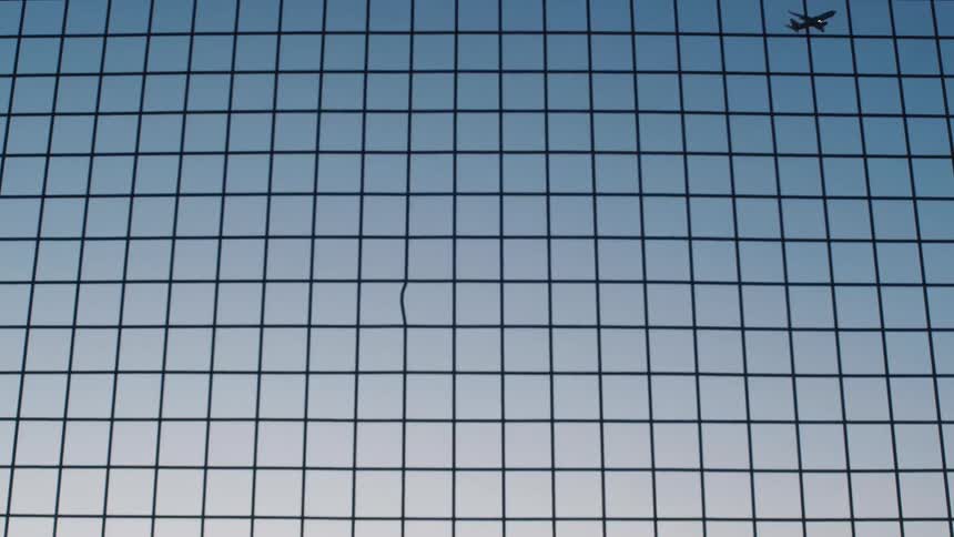 Silhouette of an airplane flying on the blue sky behind a wired fence