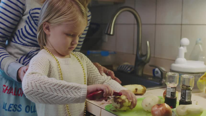 Mother teaching daughter how to peel potatoes next to the kitchen sink at home