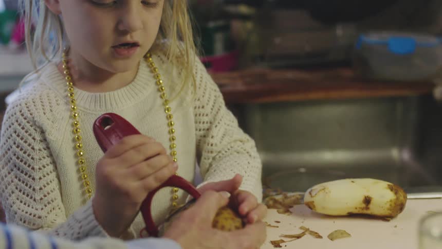 A little girl uses a peeler to peel a potato held by her grandmother who offers encouragement