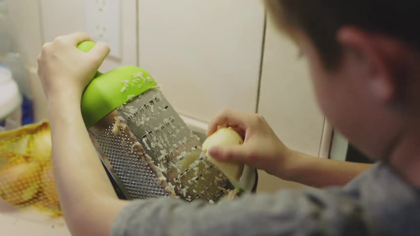 Young boy grating a raw potato inside a bowl in the kitchen at home