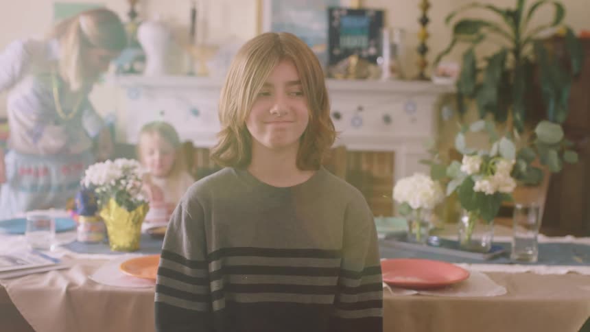 Girl stands smiling slightly as mother watches her sister eat in the background