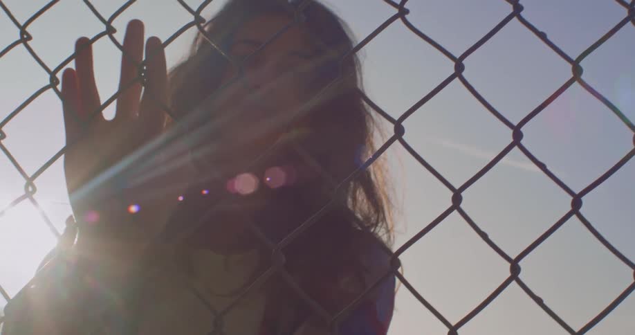 Sun back lights woman as she stands touching chain link fence
