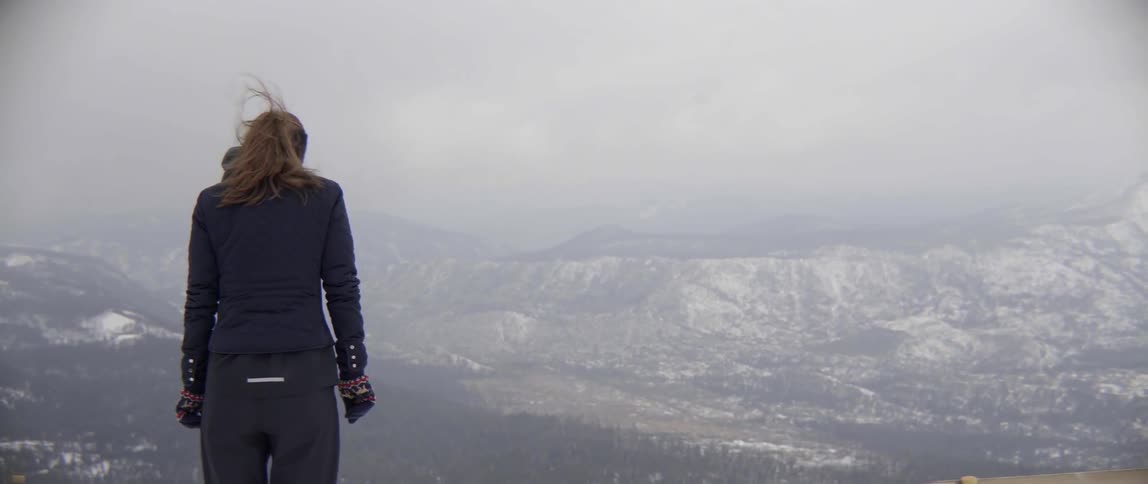 A woman stands over a snow covered mountain range on a windy day
