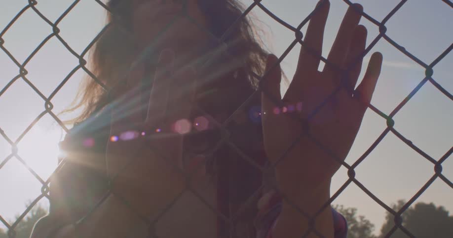 Woman touches and looks through chain link fence as she is back light by the sun