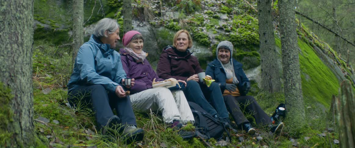 Women wearing jackets sit on ground with mugs smiling and talking with handrail following slope in the background