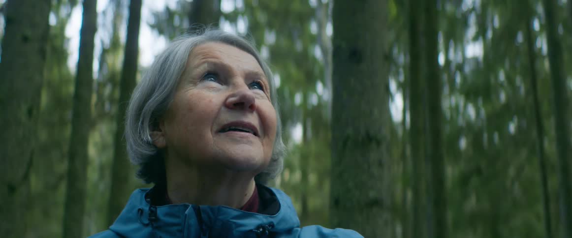An elderly woman stands in a forest and smiles as she looks around